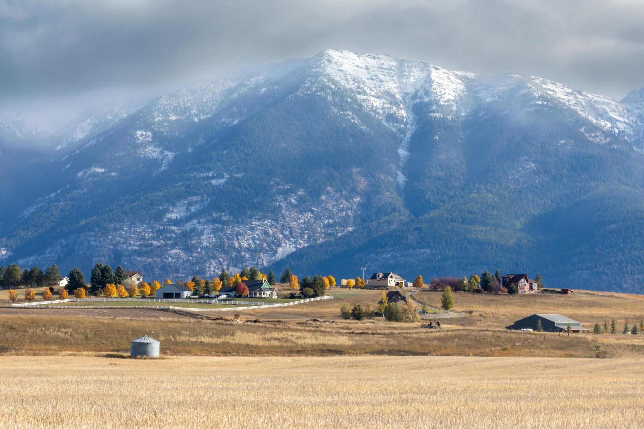 Autumnal countryside scene with snow-capped mountains. Homesteads and a grain silo are nestled in the valley. , Kalispell, Montana, USA