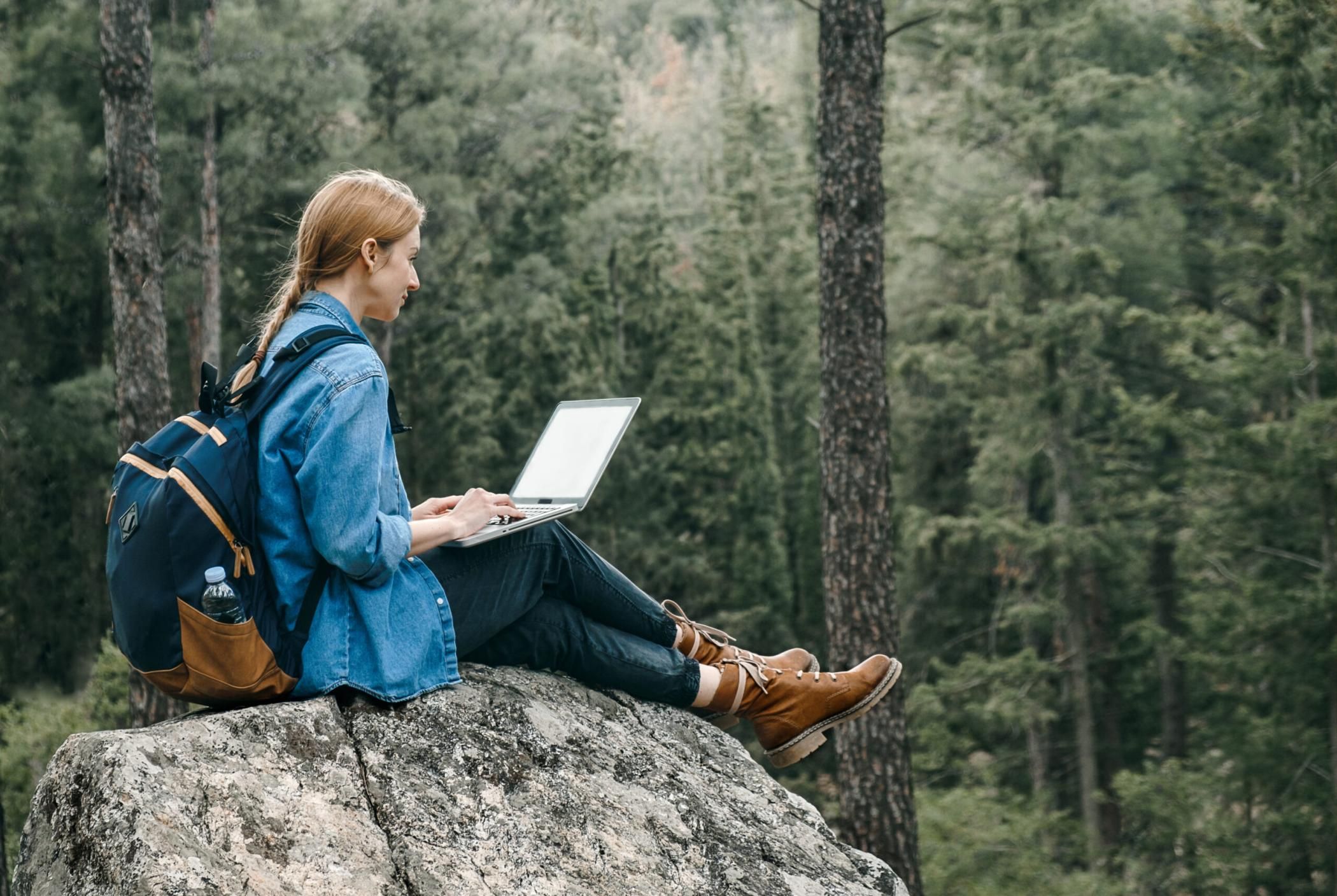 Young woman with backpack sitting on ground at forest and working on laptop . Workation concept. Freelancer work